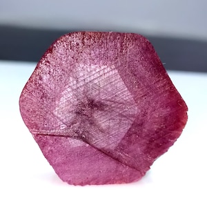 May include: A faceted, hexagonal-shaped, deep magenta gemstone. The gem has a textured surface with visible internal patterns. The stone is set against a white background, highlighting its color and form.