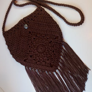 May include: A brown crocheted crossbody bag with a fringe bottom and a silver button. The bag has a long strap.