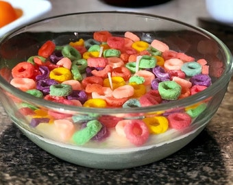 May include: A glass bowl filled with a colorful cereal-shaped candle. The candle is white with multi-colored cereal rings on top.