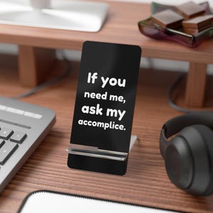 May include: A black phone stand with the text "If you need me, ask my accomplice" in white. The stand is on a wooden desk, with a keyboard, headphones, and other office supplies in the background.