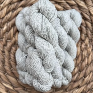 May include: A skein of light gray yarn, tightly wound and textured, rests on a woven, light brown surface. The yarn appears soft and ready for knitting or crocheting projects. The neutral color makes it versatile for various craft projects.