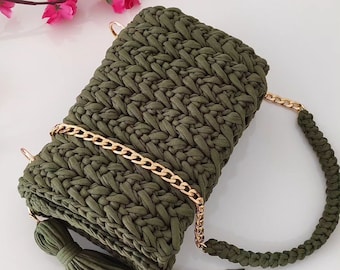 May include: An olive green crochet handbag with a gold chain strap and tassel. The bag has a woven texture and a braided handle. The gold chain is attached with gold-coloured hardware. The tassel is long and made of the same olive green material.