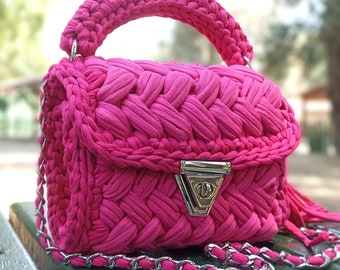 Puede incluir: Un bolso de mano de crochet de color rosa brillante con una correa de cadena plateada y un cierre plateado. El bolso tiene un borla que cuelga de la parte inferior.