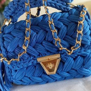 Puede incluir: Un peque&ntilde;o bolso de mano azul tejido a crochet con una correa de cadena dorada y un cierre dorado. El bolso tiene un borla que cuelga de la parte inferior.