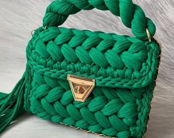 May include: A green knitted handbag with a braided handle and a gold chain strap. The bag has a gold clasp and a green tassel.