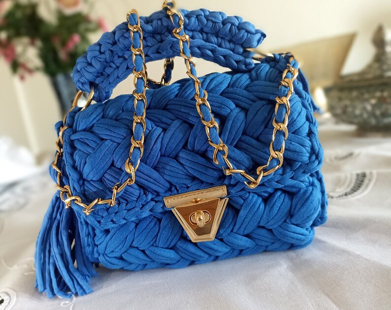 Puede incluir: Un peque&ntilde;o bolso de mano azul tejido a crochet con una correa de cadena dorada y un cierre dorado. El bolso tiene un borla que cuelga de la parte inferior.