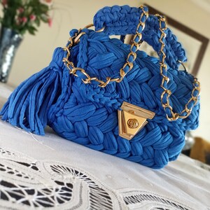 Puede incluir: Un peque&ntilde;o bolso de mano azul tejido a crochet con una correa de cadena dorada y un cierre dorado. El bolso tiene un borla en el lateral.