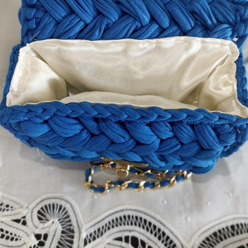 Puede incluir: Un bolso de mano de crochet azul con una correa de cadena dorada y un forro de sat&eacute;n blanco. El bolso est&aacute; abierto y el forro es visible.