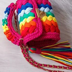 May include: A rainbow crocheted handbag with a gold chain strap and a tassel. The bag has a flap closure with a gold lock.