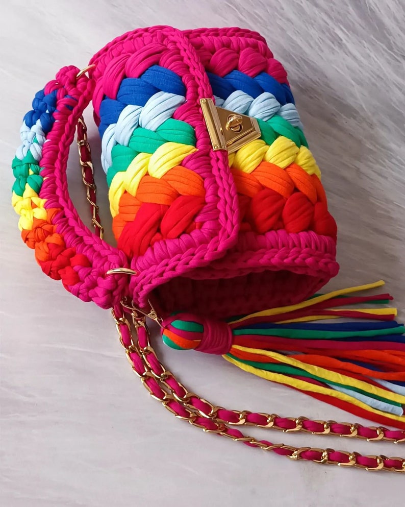 May include: A rainbow crocheted handbag with a gold chain strap and a tassel. The bag has a flap closure with a gold lock.