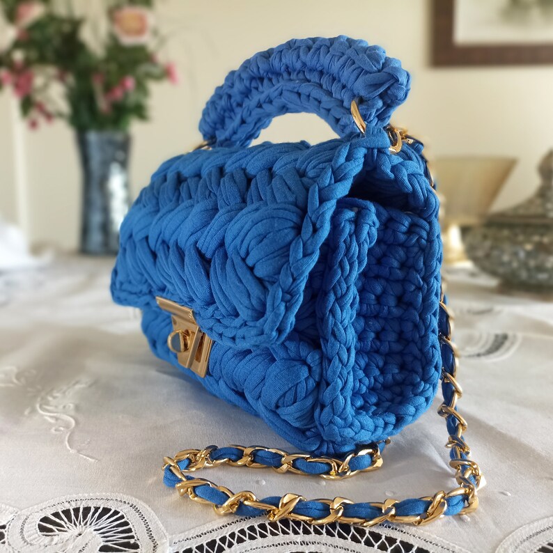 Puede incluir: Un bolso de mano de crochet azul con una correa de cadena dorada. El bolso tiene un cierre de solapa con un broche dorado.