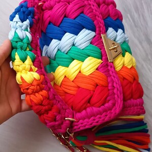 May include: A rainbow crocheted bag with a gold chain strap and a gold clasp. The bag has a fringe of rainbow colored yarn.