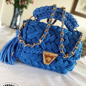 Puede incluir: Un bolso de mano azul tejido a crochet con una correa de cadena dorada y un cierre dorado. El bolso tiene un borla en el lateral.