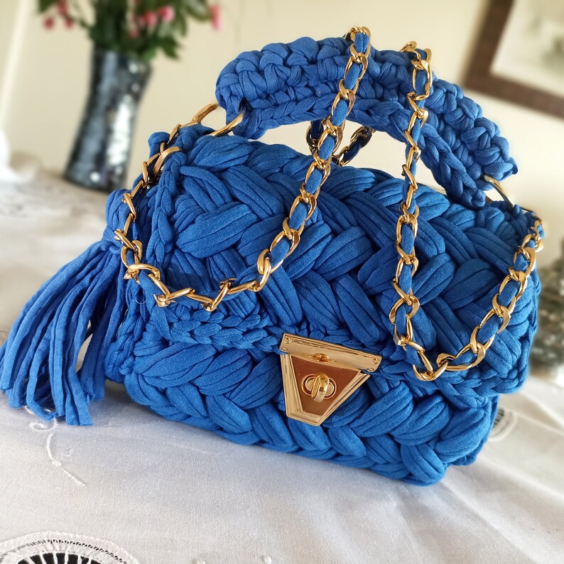 Puede incluir: Un bolso de mano azul tejido a crochet con una correa de cadena dorada y un cierre dorado. El bolso tiene un borla en el lateral.