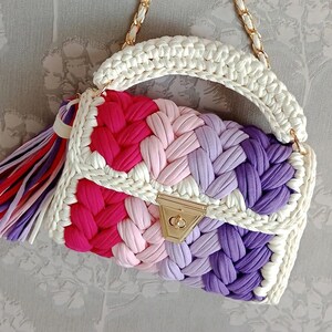 May include: A white crocheted handbag with a gold clasp. The bag has a colourful striped pattern of pink, purple, and red.