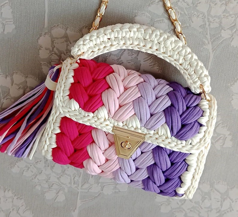May include: A white crocheted handbag with a gold clasp. The bag has a colourful striped pattern of pink, purple, and red.