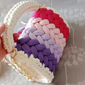 May include: A crocheted handbag with a white handle and a rainbow of colours, including pink, purple, and red. The bag has a gold chain strap.