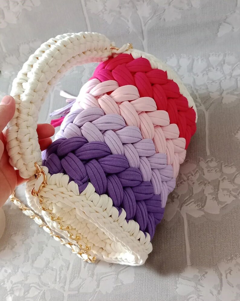 May include: A crocheted handbag with a white handle and a rainbow of colours, including pink, purple, and red. The bag has a gold chain strap.