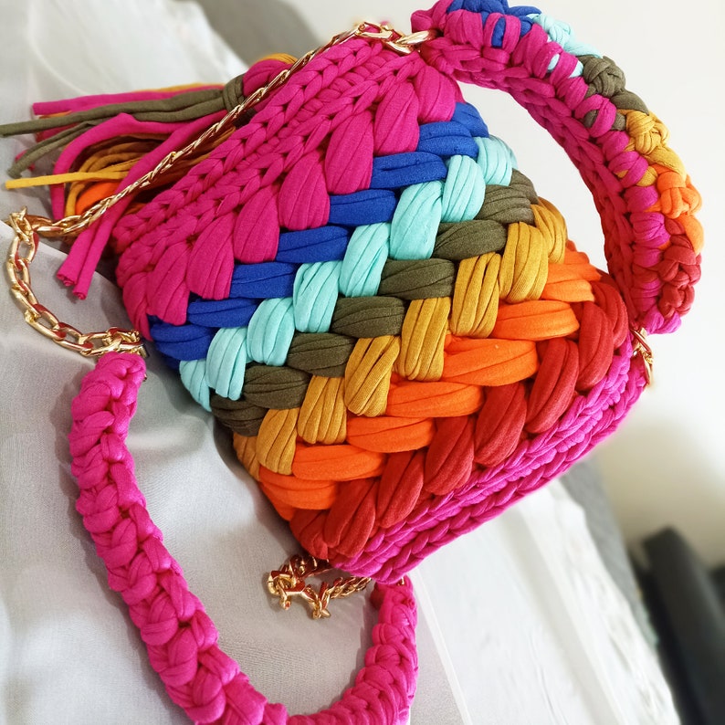 May include: A colorful crocheted bag with a rainbow pattern and a gold chain strap. The bag is made of thick yarn and has a unique, textured design.