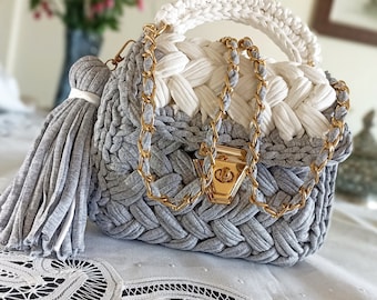 May include: A gray and white crocheted handbag with a gold chain strap and a gray tassel. The bag has a top handle and a gold clasp.