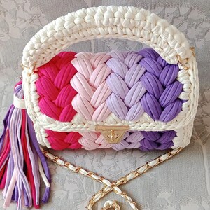 May include: A crocheted handbag with a white handle and a chain strap. The bag is made of colourful yarn in shades of pink, purple, and red. It has a gold clasp and a tassel.