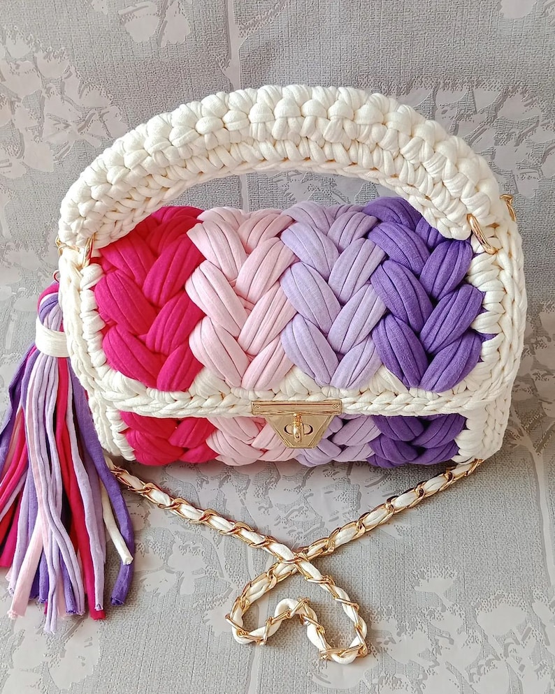 May include: A crocheted handbag with a white handle and a chain strap. The bag is made of colourful yarn in shades of pink, purple, and red. It has a gold clasp and a tassel.