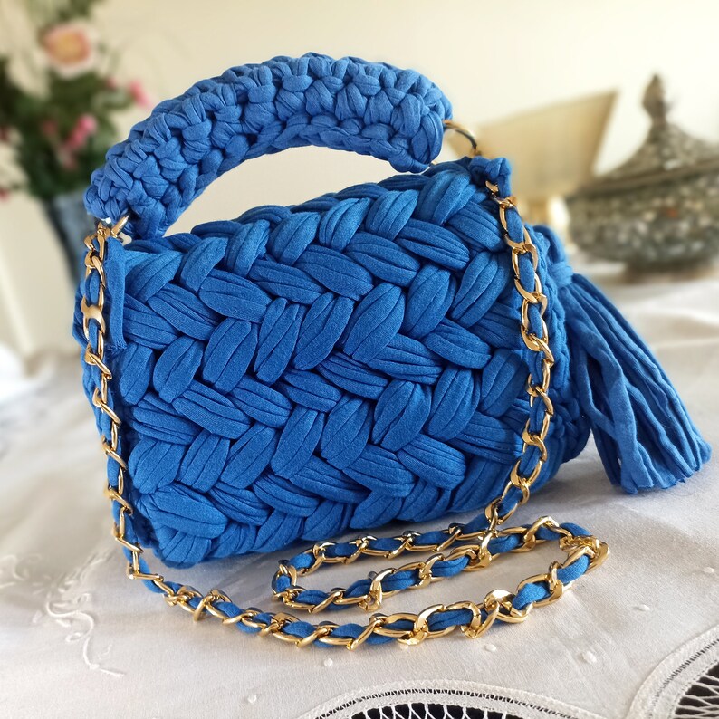 Puede incluir: Un bolso de mano de crochet azul brillante con una correa de cadena dorada y un borla. El bolso tiene una textura tejida y un asa superior.