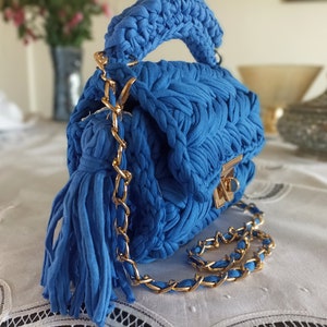Puede incluir: Un peque&ntilde;o bolso de mano tejido en azul con una correa de cadena dorada y un borla. El bolso tiene un cierre de solapa con un broche dorado.