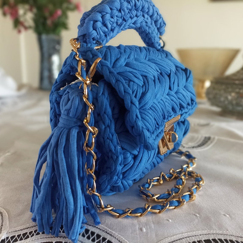 Puede incluir: Un peque&ntilde;o bolso de mano tejido en azul con una correa de cadena dorada y un borla. El bolso tiene un cierre de solapa con un broche dorado.