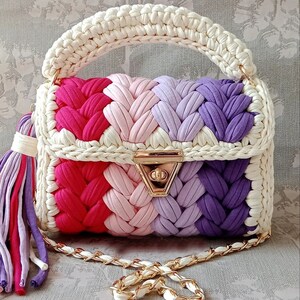 May include: A crocheted handbag with a white handle and a chain strap. The bag is made of colourful yarn in shades of pink, purple, and red. The bag has a gold clasp.