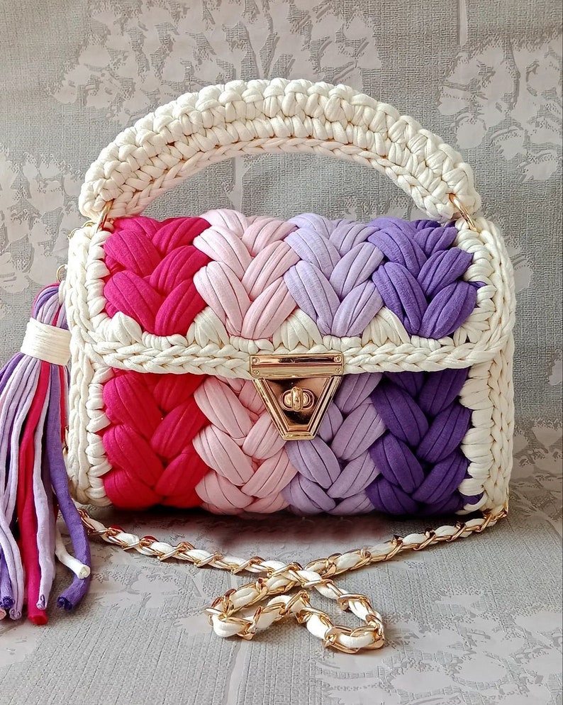 May include: A crocheted handbag with a white handle and a chain strap. The bag is made of colourful yarn in shades of pink, purple, and red. The bag has a gold clasp.