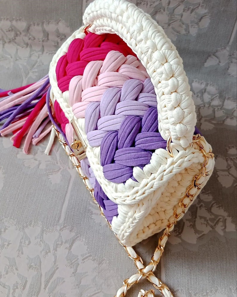 May include: A white crocheted handbag with a gold chain strap. The bag has a multi-coloured striped design with pink, purple, and white yarn.