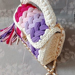 May include: A white crocheted handbag with a gold chain strap. The bag has a pink, purple, and white striped design and a tassel.