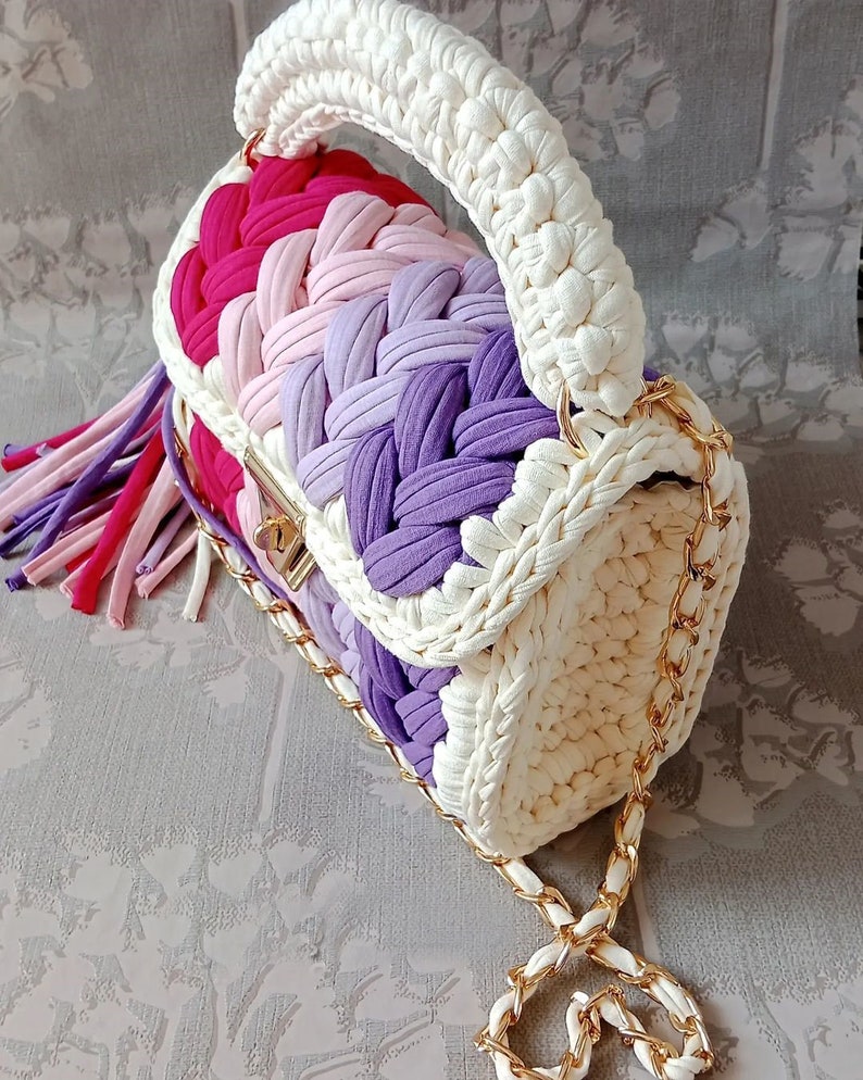 May include: A white crocheted handbag with a gold chain strap. The bag has a pink, purple, and white striped design and a tassel.