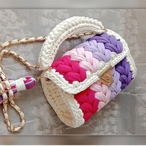May include: A white crocheted handbag with a gold chain strap. The bag has a colourful striped design with pink, purple, and white yarn.