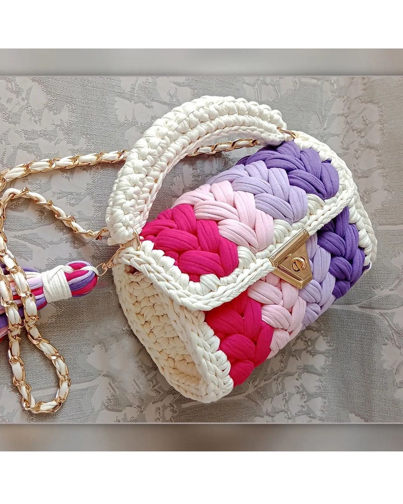 May include: A white crocheted handbag with a gold chain strap. The bag has a colourful striped design with pink, purple, and white yarn.