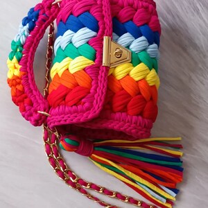 May include: A bright pink crocheted handbag with a rainbow pattern and a gold chain strap. The bag has a gold clasp and a rainbow tassel.