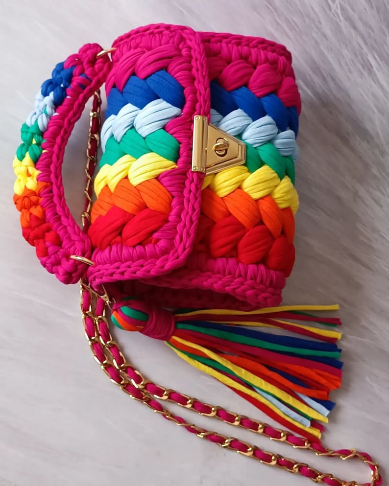 May include: A bright pink crocheted handbag with a rainbow pattern and a gold chain strap. The bag has a gold clasp and a rainbow tassel.