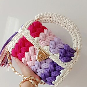 May include: A crocheted handbag with a white handle and a colourful body. The bag is made of a variety of colours, including pink, purple, and white. The bag has a gold clasp and a gold chain strap.