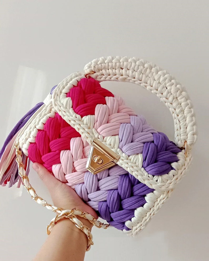 May include: A crocheted handbag with a white handle and a colourful body. The bag is made of a variety of colours, including pink, purple, and white. The bag has a gold clasp and a gold chain strap.
