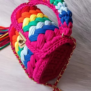 May include: A small, bright pink crocheted handbag with a rainbow-colored strap and a gold chain strap. The bag has a gold clasp.