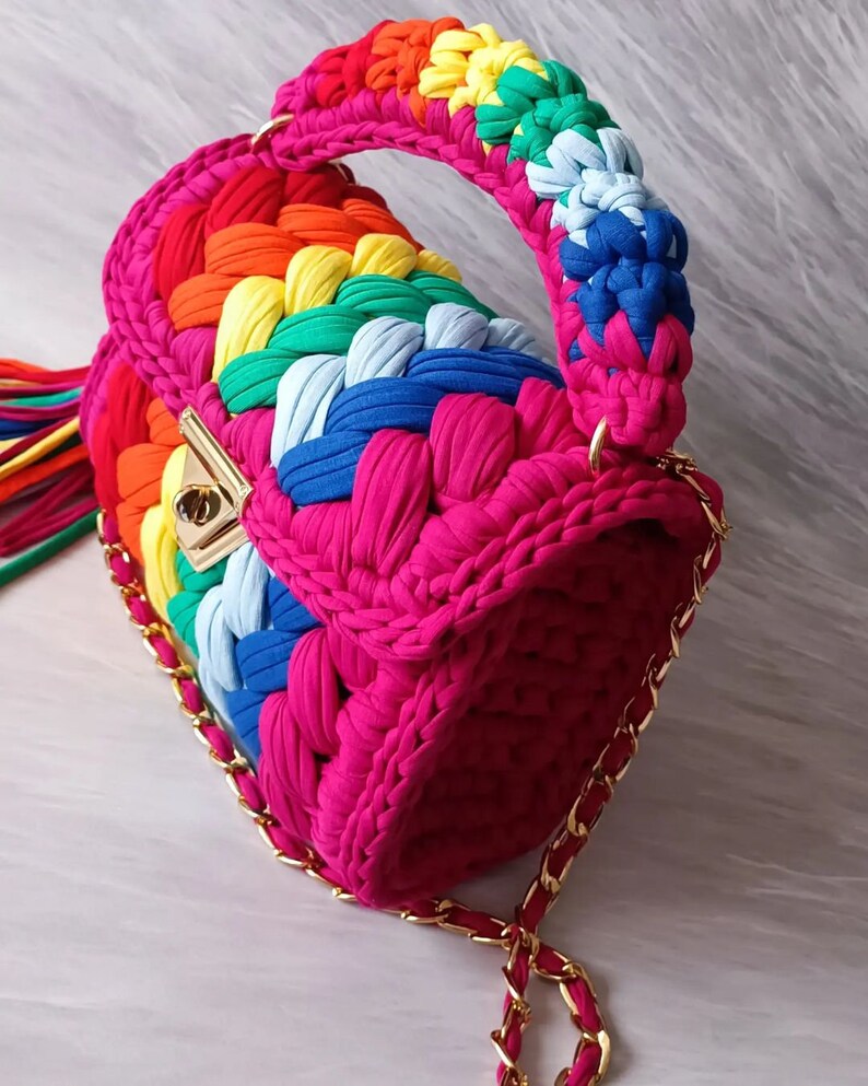 May include: A small, bright pink crocheted handbag with a rainbow-colored strap and a gold chain strap. The bag has a gold clasp.