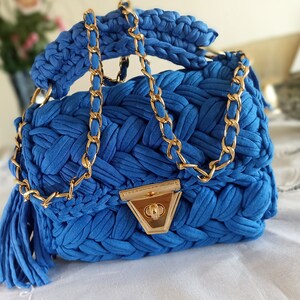 Puede incluir: Un peque&ntilde;o bolso de mano azul tejido a crochet con una correa de cadena dorada y un cierre dorado. El bolso tiene un borla que cuelga de la parte inferior.