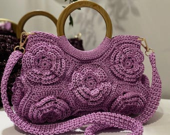 May include: A lavender crochet handbag with a floral design. The bag features a wooden handle and a detachable shoulder strap. The bag is approximately 10 inches wide and 8 inches tall.