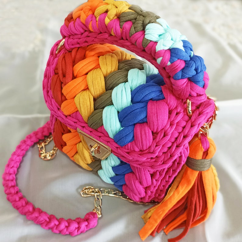 May include: A colorful crocheted handbag with a gold clasp and a long strap. The bag is made of different colored yarn, including pink, orange, blue, green, and turquoise.