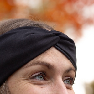 May include: A black turban-style headband made of soft fabric. The headband is knotted at the top and worn across the forehead. The background is blurred, with warm orange and white tones.