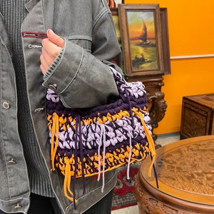 May include: A handmade crochet bag in shades of purple, orange, and lavender. The bag features a textured design with fringe detailing. The bag is held by a person wearing a gray denim jacket.