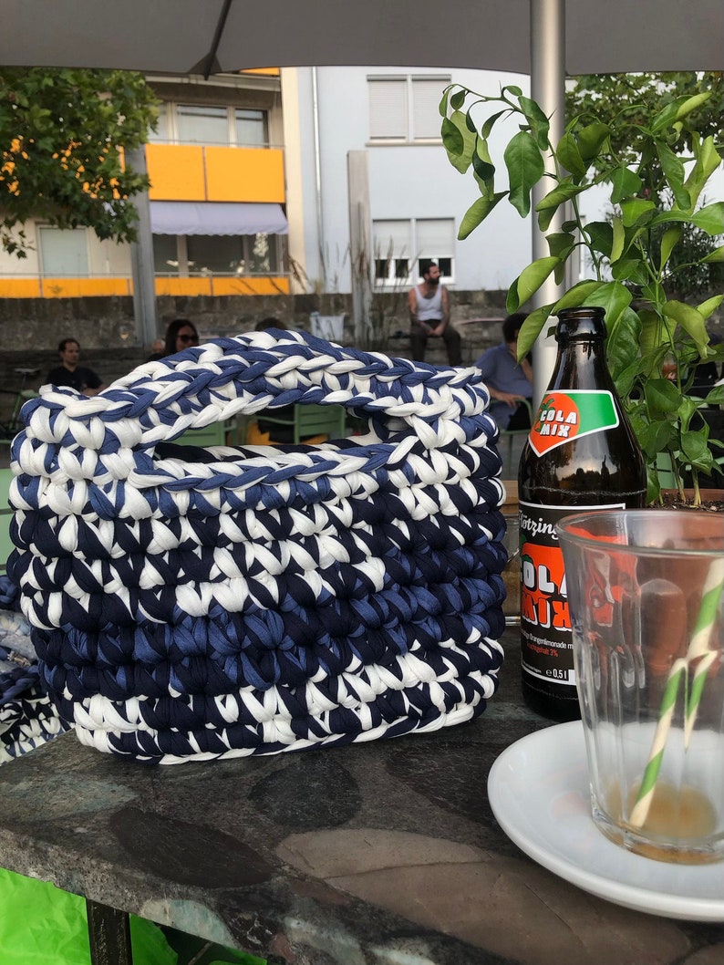 May include: A handmade, rectangular basket with a handle, crafted from thick, woven fabric in alternating shades of navy blue and white. The basket sits on a stone surface, with a bottle of Cola Mix and a glass nearby.