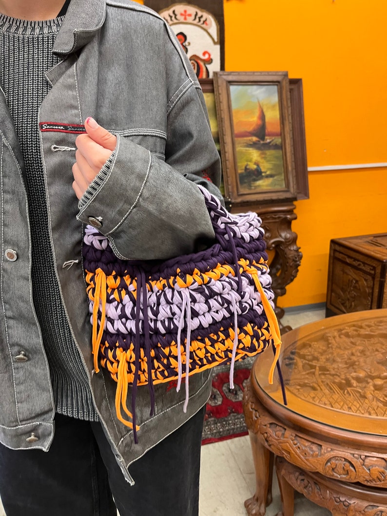 May include: A handmade crochet bag in shades of purple, orange, and lavender. The bag has a rectangular shape with a fringed edge and a short handle. The bag is held by a person wearing a gray denim jacket.