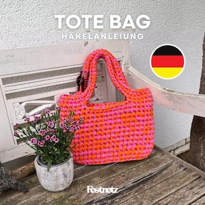 Puede incluir: Un bolso tote hecho a mano con hilo rosa y naranja vibrante descansa sobre un banco de madera. El bolso presenta un diseño de ganchillo resistente con asas a juego. Las palabras "TOTE BAG" y "HÄKELANLEITUNG" se muestran en la parte superior.